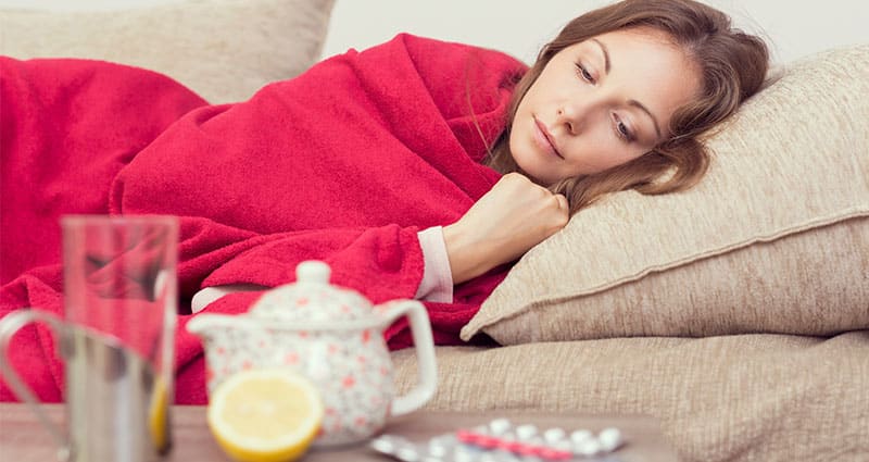 Woman resting on sofa with medication and teapot nearby.