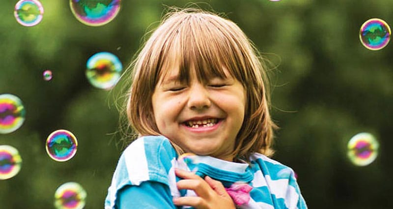 A little girl smiles with her eyes closed as soap bubbles float in the air around her.