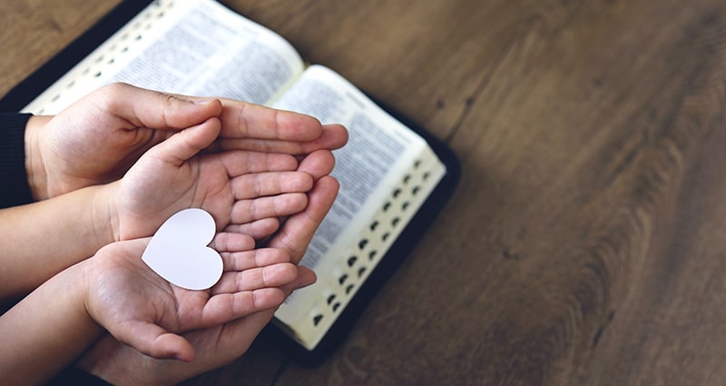 paper heart in child's hands being held by parent's hands resting on top of open Bible