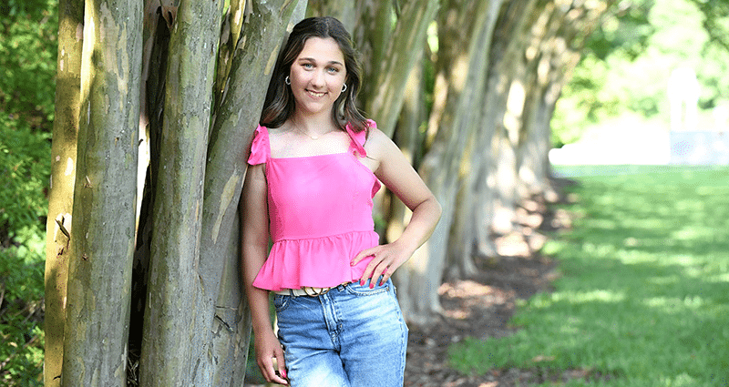 Lauren, smiling teenager in pink shirt and jeans by a tree