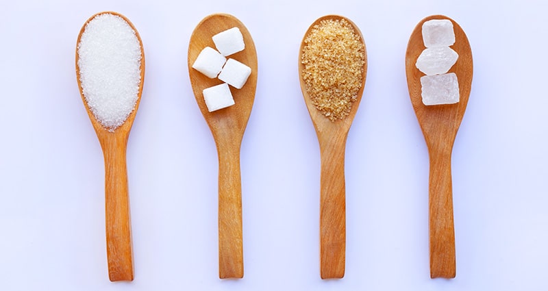 spoons with different types of sugar on light purple background