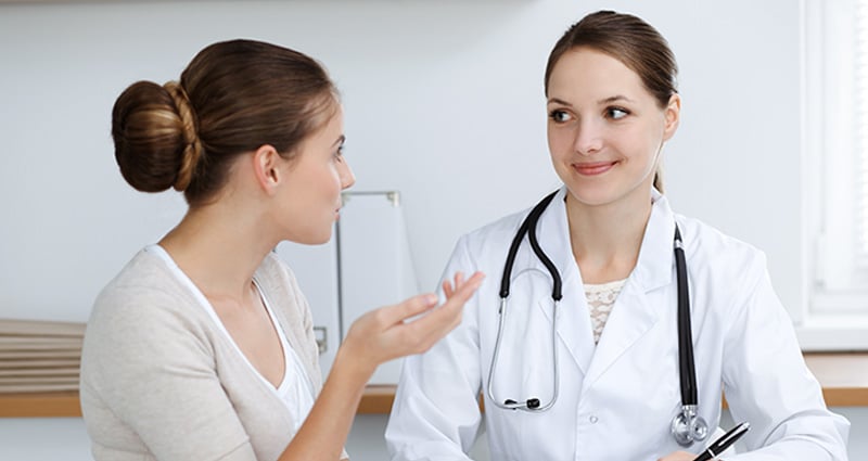 A young woman with her hair in a bun gestures as if speaking. Another woman wearing a white coat and stethoscope listens attentively.