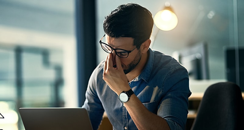 A man wearing glasses looks at a laptop. He is holding the bridge of his nose as if he has a headache.