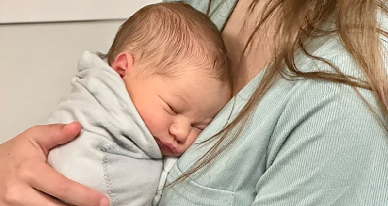 In a close-up image, a baby rests against a woman's chest. The woman is wearing a green shirt, while the baby is swaddled snugly.