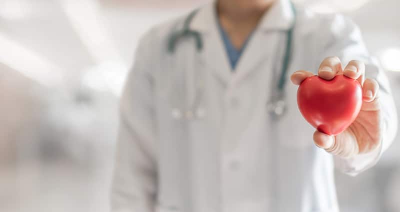 A man wearing a white medical coat holds a red, heart-shaped toy out toward the camera.