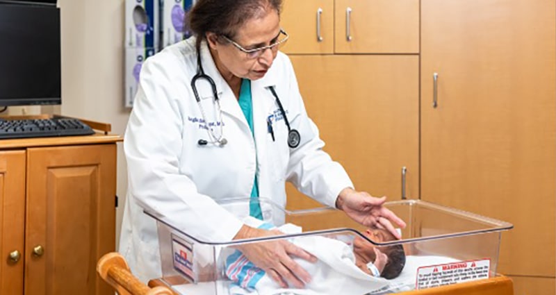 A woman wearing a white coat with a stethoscope around her neck tends to an infant. There is a loving expression on her face.