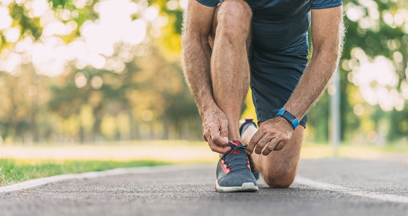 In this close-up image, a man is tying his sneaker. He's kneeling on a concrete surface with trees and sunlight behind him.