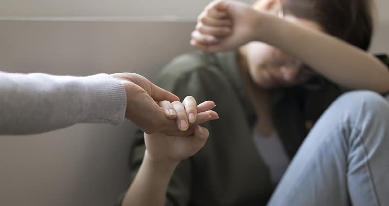 A young man sits with his back against a wall. He has one arm over his eyes. From outside the image, an adult reaches out toward him. He grasps her hand.