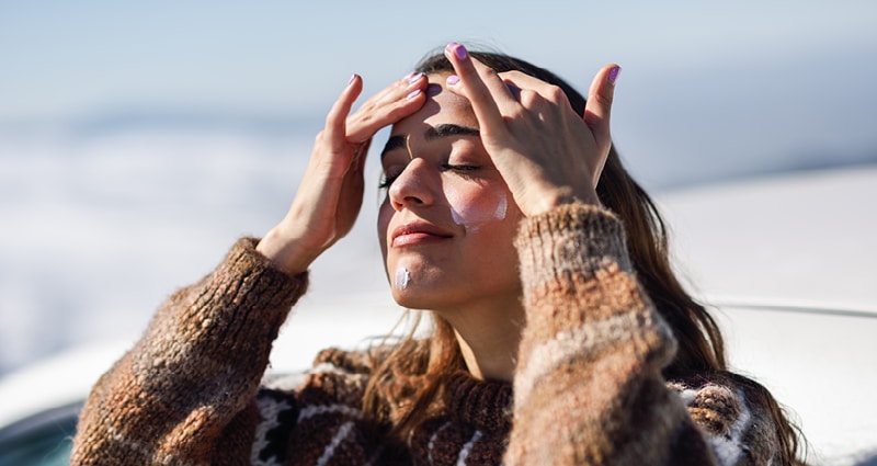 A woman wearing a brown sweater stands in front of a body of water. She is applying sunscreen to her forehead.