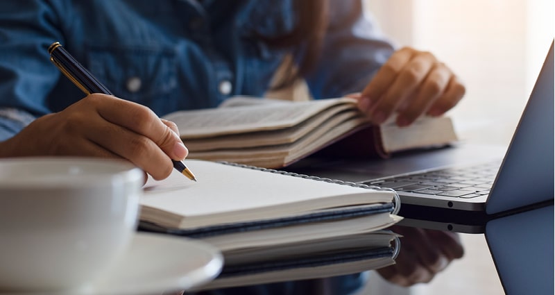 In this close-up photo, a person is reading from one notebook and making notes in another. We see only their hands and a glimpse of their blue shirt. A laptop is open in front of them.