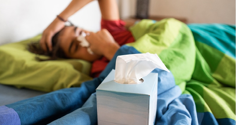 A young person lies in bed under a blue and green blanket. She is blowing her nose. A box of tissues is in the foreground.