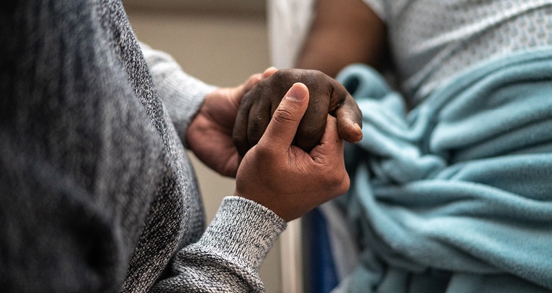 In a close-up image, a man is lying on a hospital bed with a blue blanket over him. Another man holds the first man's hand reassuringly in both of his.