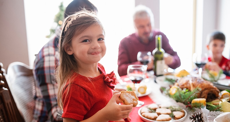 A family of four enjoys a holiday meal. In the foreground, a little girl in a red dress grins at the camera. She is holding a cookie.