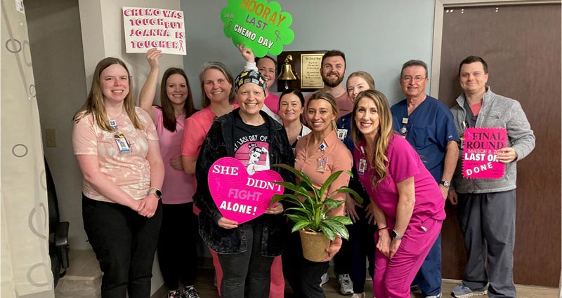 In this group photo, people wearing medical name badges or uniforms cluster around a woman holding a large, pink cardboard heart. They are smiling. Others hold posters celebrating her last day of chemotherapy.