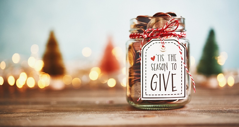 In the background on a wooden surface, Christmas lights glisten. In the foreground, a jar of coins has a label reading, "Tis the Season to Give."