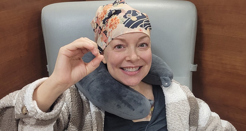 A woman wearing a bright floral bandana on her head makes an "O" sign with her hand. She is smiling.