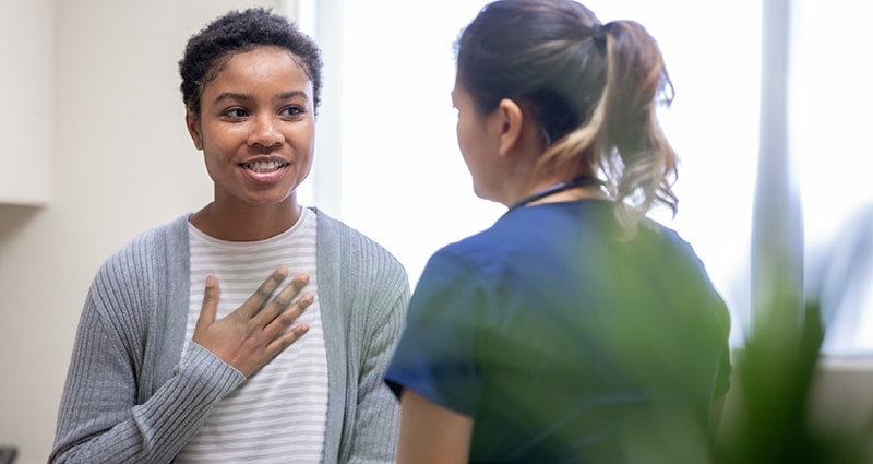 A young woman smiles and holds her hand over her heart as she talks with a medical provider. Facing away from the camera, the medical provider appears to be listening sympathetically.