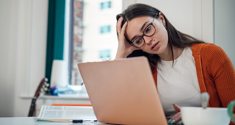 A young woman wearing glasses leans her head on her hand and looks at the screen of a laptop computer.