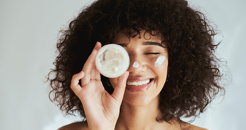 A young woman smiles radiantly at the camera. She is holding a container of skincare product in front of one eye. The same skincare product is dabbed on her cheek and nose.