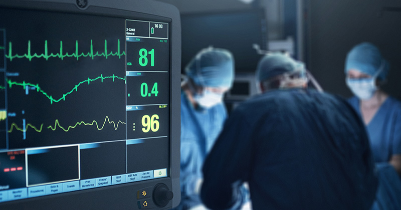 A heart monitor is in the foreground while surgeons are performing surgery in the background